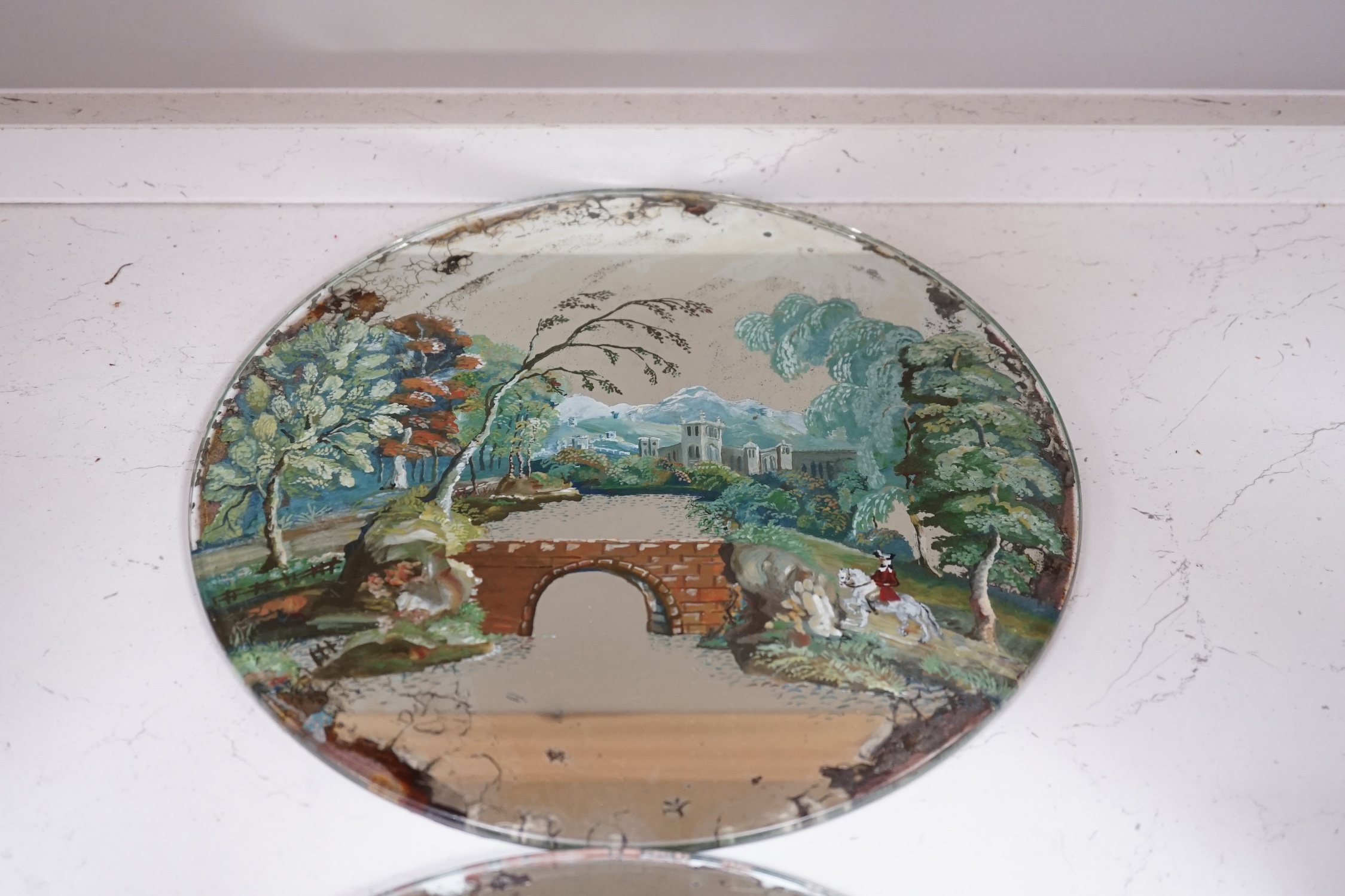 Three reverse painted mirrored glass place mats including a pair depicting landscapes with figures on horseback, largest 30cm diameter. Condition - fair